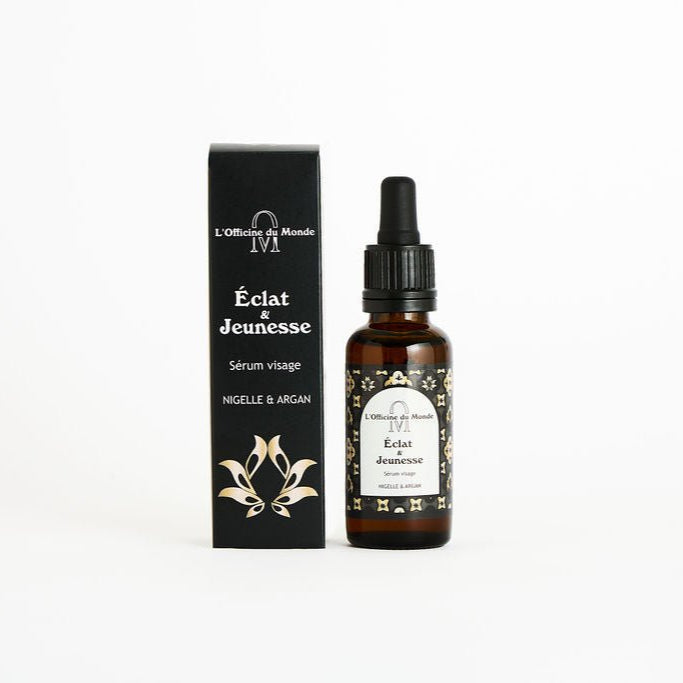 Flacon en verre brun du sérum visage « Éclat &amp; Jeunesse » de L’Officine du Monde, avec pipette noire et étiquette noire et dorée, posé à côté de sa boîte noire assortie sur fond blanc.